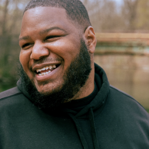Kirwyn Sutherland, a Black poet with a close beard is looking over his shoulder and smiling. He wears a black sweatshirt and it looks to be a fall day outdoors.